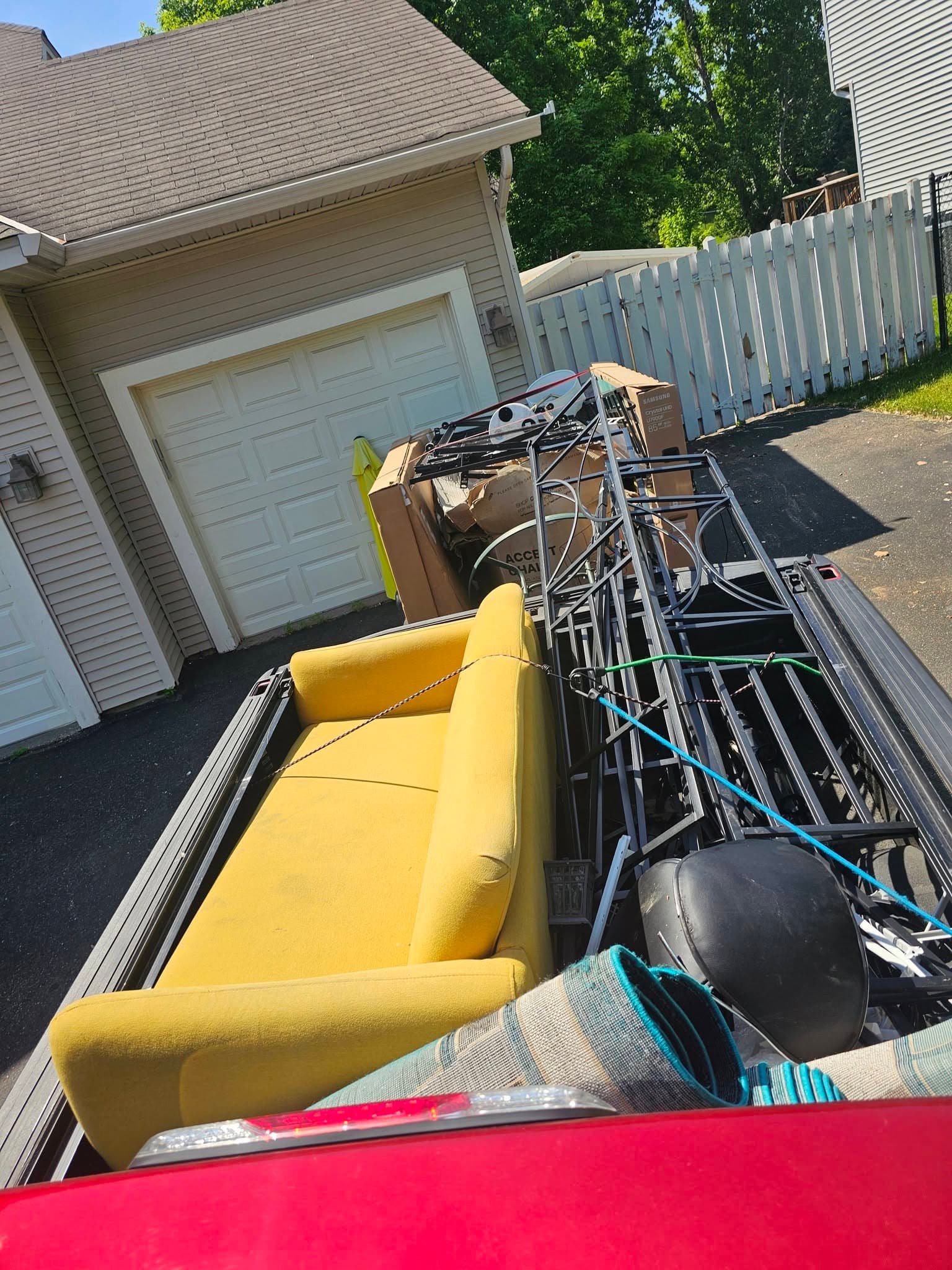 Open utility trailer packed with a yellow sofa, metal frames, boxes, and mixed household items beside a garage.
