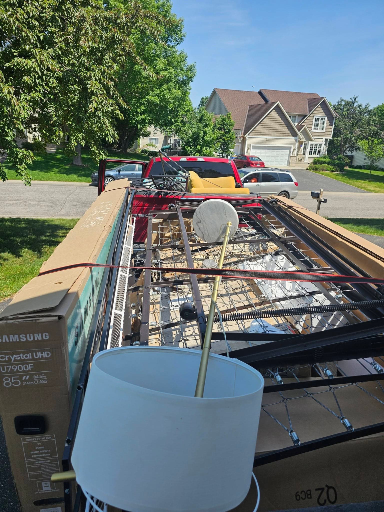 Utility trailer and red pickup loaded with metal bed frames, boxes, a lamp, and mixed household items for hauling.